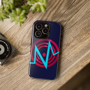 May include: A navy blue phone case featuring a red and blue graphic design. The design includes concentric circles and a stylized blue line. The phone case is on a wooden surface, with a camera and plant in the background.