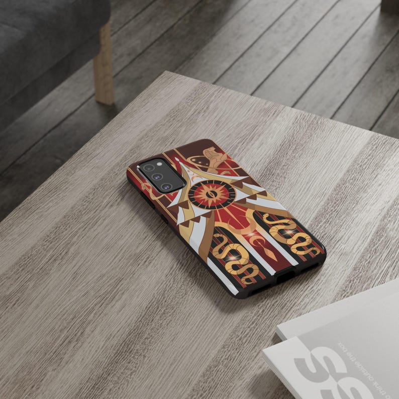 May include: A smartphone case with a geometric design featuring a central sunburst motif in red, gold, and white. The case has a dark brown border and a pattern of stylized snakes. The case is lying on a wooden table.