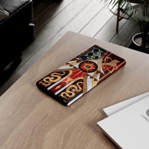 May include: A phone case with a geometric design in red, gold, white, and black. The case features a central sunburst pattern and stylized snake and archer motifs. The case is on a light brown wooden surface.