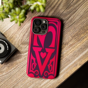 Op de afbeelding: Een rode en paarse telefoonhoes met het woord "LOVE" in een vetgedrukte, gestileerde lettertype. Het ontwerp bevat een hart en decoratieve krullen. De hoes ligt op een houten oppervlak, met een camera en groen in de achtergrond.