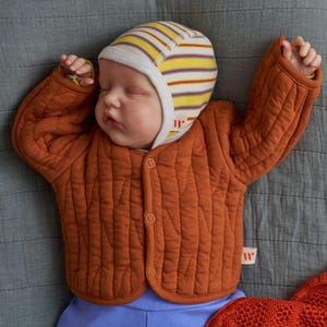 Könnte beinhalten: Ein Baby trägt eine braune gesteppte Jacke mit einer gelb-braunen gestreiften Mütze. Das Baby liegt auf einer grauen Decke.