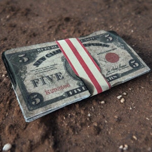 K&ouml;nnte beinhalten: Ein Stapel von F&uuml;nf-Dollar-Scheinen, die mit einem rot-wei&szlig;-blauen Gummiband zusammengehalten werden. Die Scheine sind alt und abgenutzt, und das Gummiband ist leicht verkohlt.