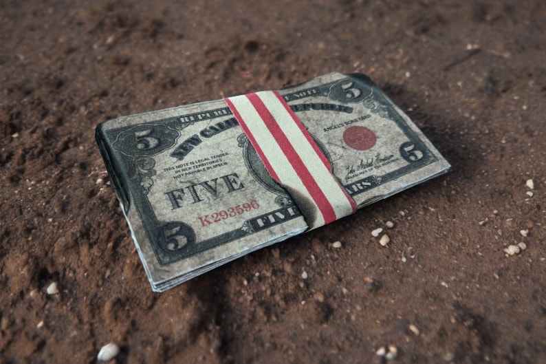 K&ouml;nnte beinhalten: Ein Stapel von F&uuml;nf-Dollar-Scheinen, die mit einem rot-wei&szlig;-blauen Gummiband zusammengehalten werden. Die Scheine sind alt und abgenutzt, und das Gummiband ist leicht verkohlt.