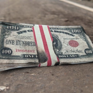 K&ouml;nnte beinhalten: Ein Stapel alter, abgenutzter 100-Dollar-Scheine mit einem rot-wei&szlig;-blauen Band darum. Die Scheine liegen auf einem Feldweg.