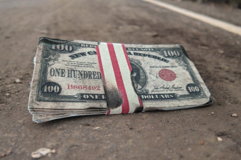 K&ouml;nnte beinhalten: Ein Stapel alter, abgenutzter 100-Dollar-Scheine mit einem rot-wei&szlig;-blauen Band darum. Die Scheine liegen auf einem Feldweg.
