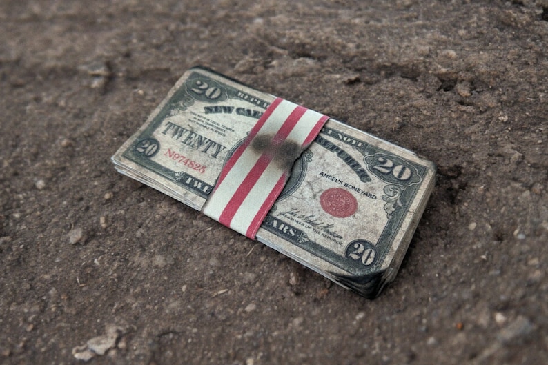 K&ouml;nnte beinhalten: Ein Stapel von Zwanzig-Dollar-Scheinen mit einem rot-wei&szlig;-blauen Band darum. Die Scheine sind leicht verkohlt und scheinen verbrannt zu sein.