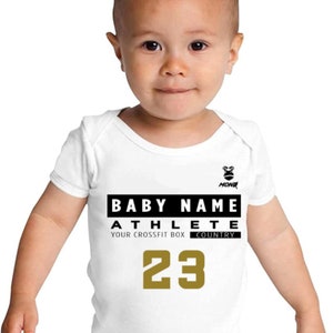 Puede incluir: Un body blanco para bebé con texto negro que dice "BABY NAME ATHLETE YOUR CROSSFIT BOX COUNTRY 23". El body tiene un pequeño logotipo blanco y negro en la esquina superior derecha.