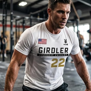 Könnte beinhalten: Ein weißes kurzärmeliges T-Shirt mit einer kleinen amerikanischen Flagge, einem schwarzen Logo und dem Text GIRDLER CROSSFIT CALGARY in schwarzer Schrift. Die Zahl 22 ist in großer goldener Schrift auf der Vorderseite des T-Shirts mit Rundhalsausschnitt gedruckt.