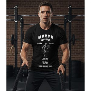 Peut inclure: T-shirt noir avec l'inscription blanche "MURPH HERO WOD NEVER FORGET". En dessous, un graphique d'un coureur devant un drapeau américain. Le t-shirt répertorie également les instructions d'entraînement. La personne sur l'image tient des cordes.