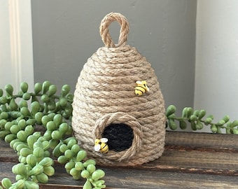 Puede incluir: Adorno decorativo en forma de colmena hecho de cuerda enrollada. La colmena tiene una abertura circular y un lazo en la parte superior. Dos pequeñas figuras de abeja amarillas y negras están unidas al exterior. El adorno se exhibe sobre una superficie de madera.
