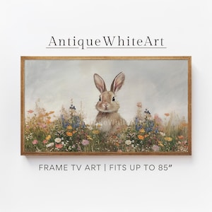 Op de afbeelding: Een ingelijste print van een bruin konijn dat in een veld met kleurrijke wilde bloemen zit. Het konijn kijkt recht naar de kijker. De print is in een houten lijst en heeft de tekst "Antique White Art" bovenaan en "FRAME TV ART | FITS UP TO 85" onderaan.