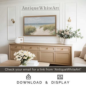 May include: A framed seascape painting of a sandy beach with green grass and a blue ocean in the background. The painting is hanging on a white wall above a light brown dresser with six drawers. The dresser is in a living room with a white couch and a vase of white flowers on the dresser.