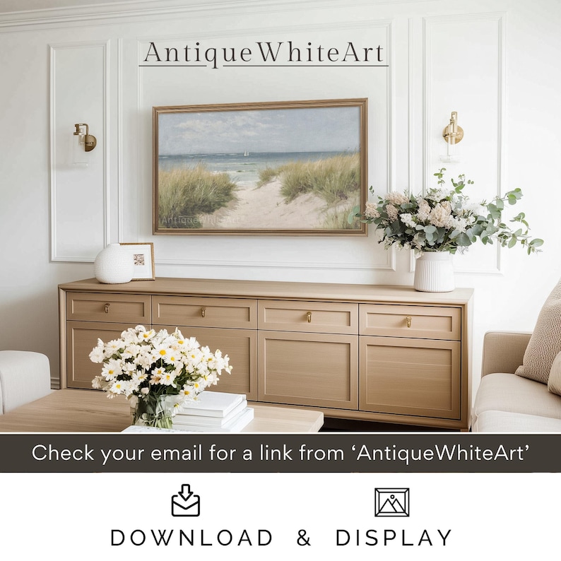 May include: A framed seascape painting of a sandy beach with green grass and a blue ocean in the background. The painting is hanging on a white wall above a light brown dresser with six drawers. The dresser is in a living room with a white couch and a vase of white flowers on the dresser.