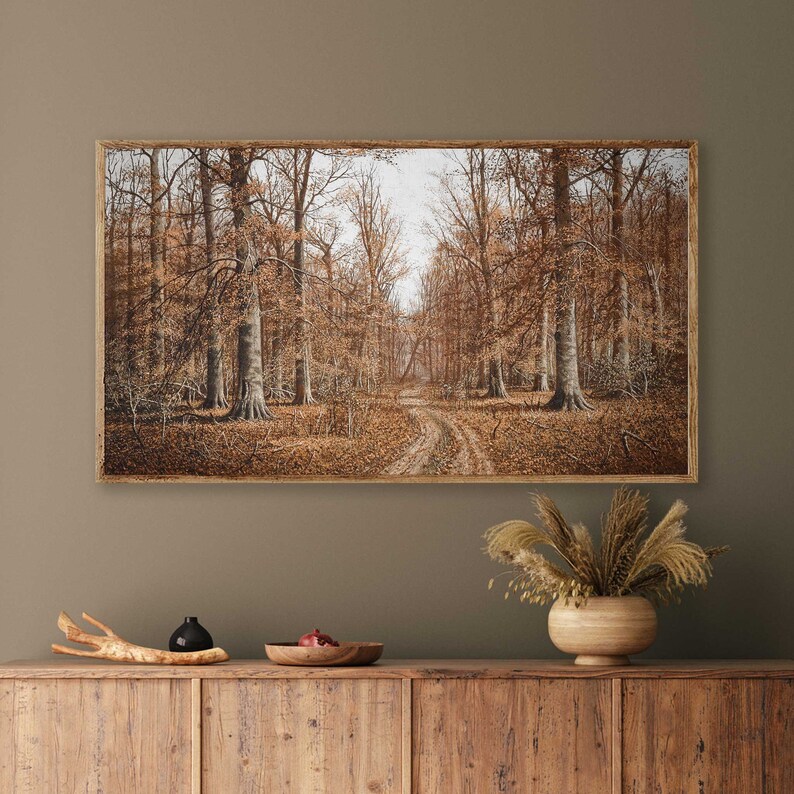 May include: A framed landscape painting of a forest scene in autumn. The artwork depicts a dirt path winding through a forest of tall trees with brown leaves. The painting is in a wooden frame and hangs on a green wall.
