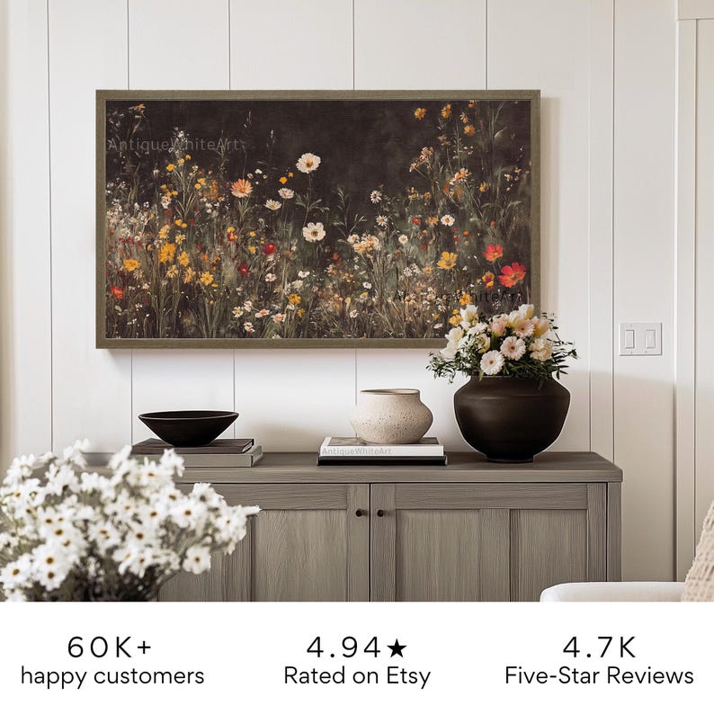 May include: A framed print of a dark floral painting featuring a variety of flowers in shades of pink, red, yellow, and white. The print is hanging on a white wall above a wooden cabinet with a vase of flowers and a bowl.