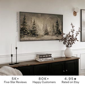 May include: A framed landscape art print depicting a winter forest scene with snow-covered trees in shades of gray and beige. The artwork hangs above a black cabinet with a wooden top, complemented by decorative candles and a vase with branches.