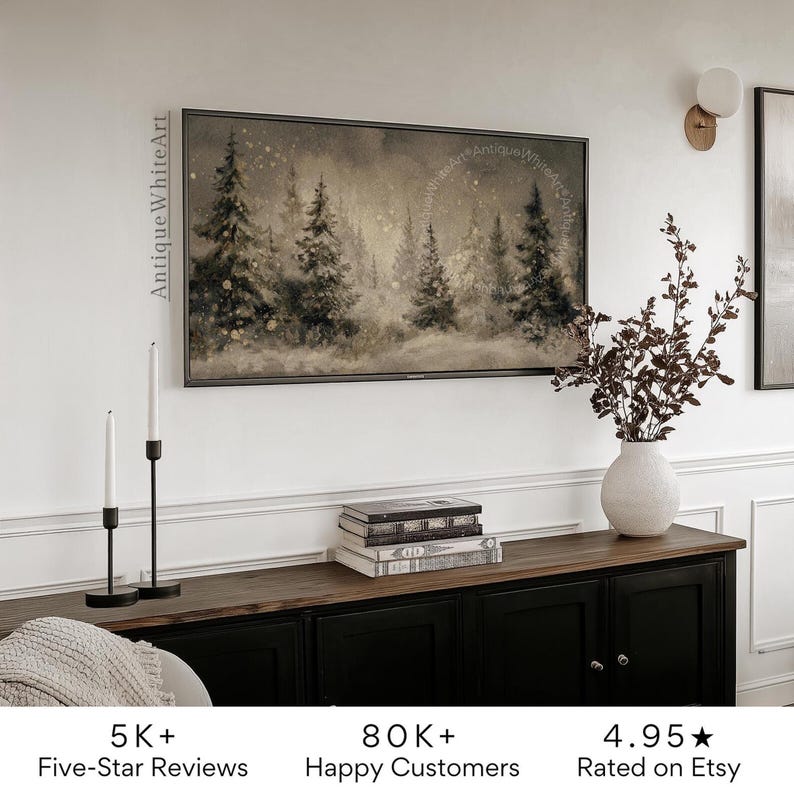 May include: A framed landscape art print depicting a winter forest scene with snow-covered trees in shades of gray and beige. The artwork hangs above a black cabinet with a wooden top, complemented by decorative candles and a vase with branches.