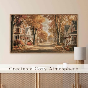 May include: A framed print of an autumnal street scene, featuring houses with porches, trees with golden leaves, and pumpkins. The text at the bottom reads "Creates a Cozy Atmosphere."