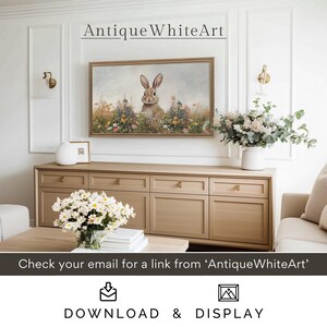 Op de afbeelding: Een digitale download van een schilderij met een wit konijn dat in een veld met wilde bloemen zit. Het schilderij is ingelijst en hangt aan een muur boven een houten dressoir. De tekst "Antique WhiteArt" wordt boven het schilderij weergegeven.