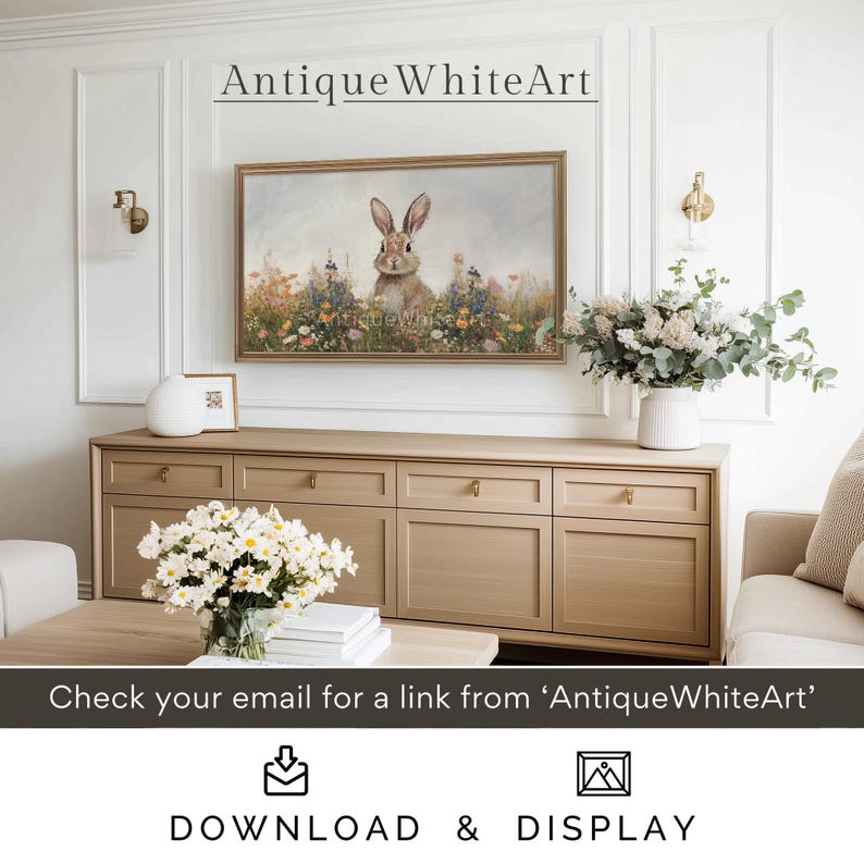 Op de afbeelding: Een digitale download van een schilderij met een wit konijn dat in een veld met wilde bloemen zit. Het schilderij is ingelijst en hangt aan een muur boven een houten dressoir. De tekst "Antique WhiteArt" wordt boven het schilderij weergegeven.