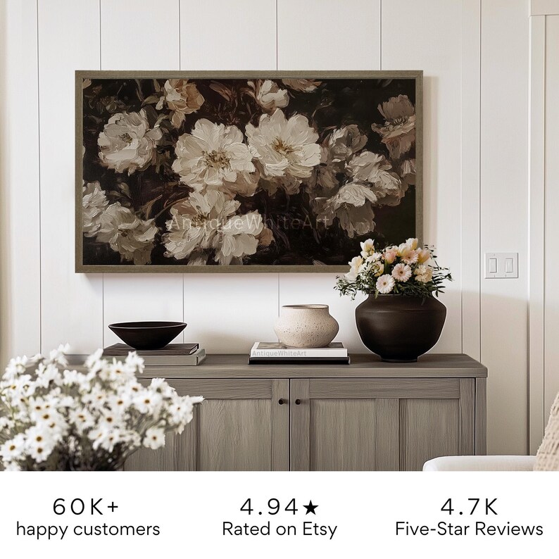 May include: A framed painting of white flowers with a dark background. The painting is hanging on a white wall above a wooden cabinet. The cabinet has two doors and two drawers. There are flowers in a vase on the cabinet. The image also includes text that says "60K+ happy customers", "4.94 Rated on Etsy", and "4.7K Five-Star Reviews".