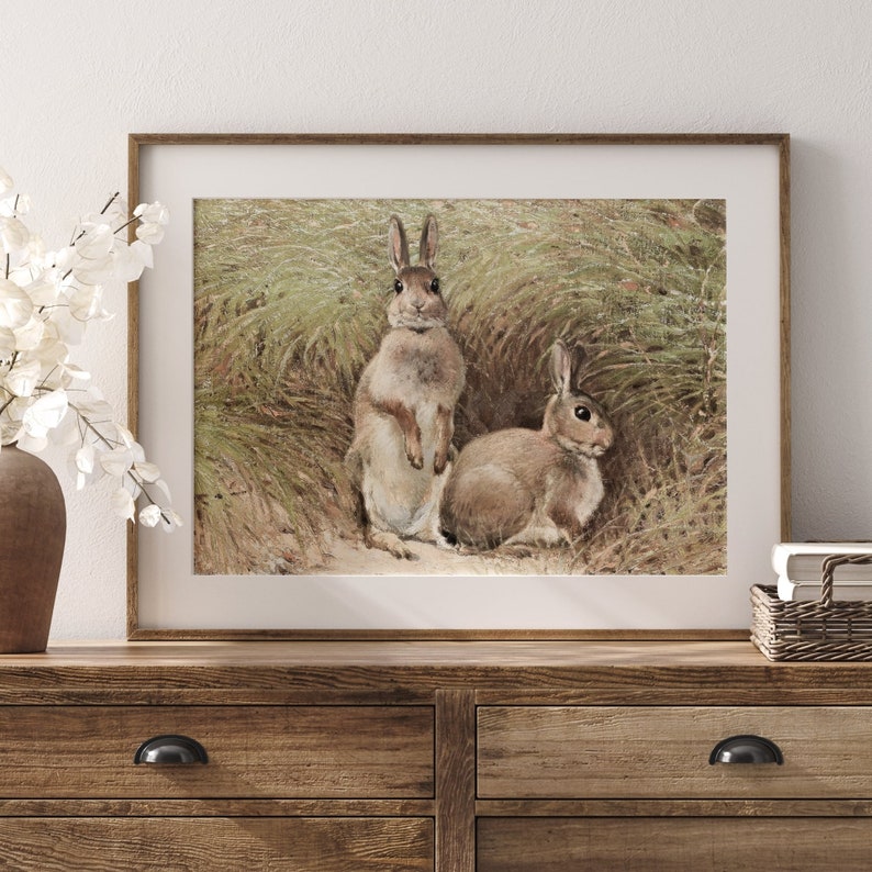 Peut inclure: Une illustration vintage de deux lapins bruns dans un champ d'herbe verte. Le lapin de gauche est debout, les pattes crois&eacute;es, tandis que le lapin de droite est assis. L'illustration est encadr&eacute;e dans un cadre en bois brun clair.