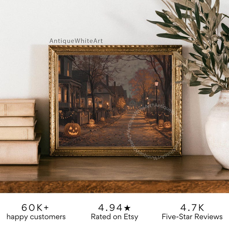 May include: A framed print of a Halloween scene with a street lined with houses, lit by streetlights and jack-o'-lanterns. The print is in a gold frame and is displayed on a wooden surface. The text "AntiqueWhiteArt" is visible.