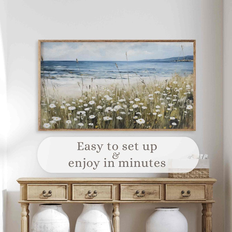 May include: A framed landscape painting depicts a coastal scene with a beach, ocean, and sky. The artwork features white flowers and tall grass in the foreground. The text "Easy to set up & enjoy in minutes" is below the painting.