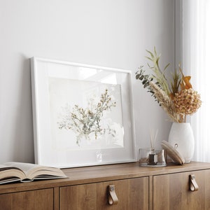 K&ouml;nnte beinhalten: Ein wei&szlig; gerahmter Druck einer botanischen Illustration eines Trockenblumenarrangements auf einem Holzregal mit einer Vase mit Trockenblumen und einer Diffuserflasche.