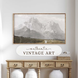 May include: A vintage art print depicting a mountain range with snow-capped peaks and a winding path leading through a grassy field. The print is framed in a wooden frame and hangs on a white wall above a wooden console table.