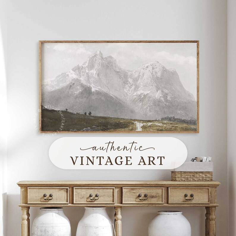 May include: A vintage art print depicting a mountain range with snow-capped peaks and a winding path leading through a grassy field. The print is framed in a wooden frame and hangs on a white wall above a wooden console table.