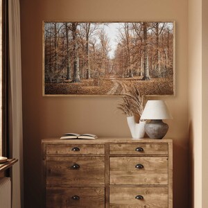 May include: A framed landscape painting depicts a forest scene with a dirt path winding through trees. The artwork is displayed above a wooden dresser with a lamp, vase, and open book. The color palette is dominated by warm browns and tans.