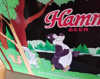 May include: A vintage Hamm's Beer sign featuring a black and white bear, a rabbit, and a squirrel running through a forest. The sign reads "Hamm's Beer" in red lettering and "Refreshing as the Land of Sky Blue Waters" in black lettering.