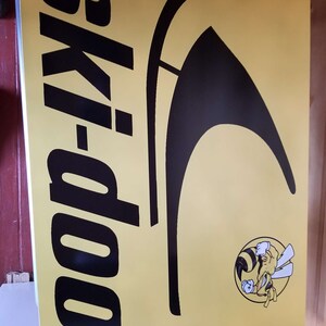 Puede incluir: Un letrero amarillo con las palabras "ski-doo" en letras negras grandes. Tambi&eacute;n hay un gr&aacute;fico negro y un logotipo de abeja de dibujos animados. El letrero es probablemente para una moto de nieve o deportes de invierno.