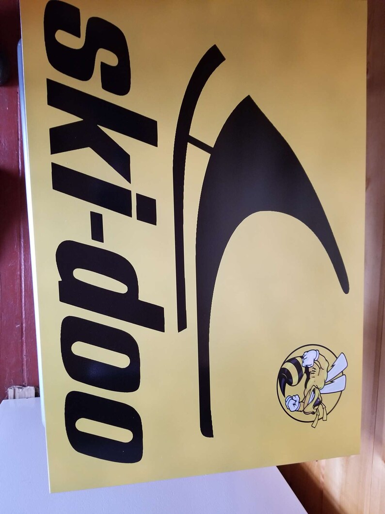 Puede incluir: Un letrero amarillo con las palabras "ski-doo" en letras negras grandes. Tambi&eacute;n hay un gr&aacute;fico negro y un logotipo de abeja de dibujos animados. El letrero es probablemente para una moto de nieve o deportes de invierno.