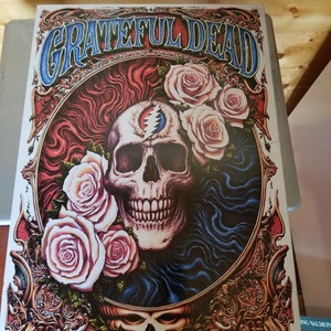 May include: A vintage-style Grateful Dead poster featuring a skull with a lightning bolt in its forehead, surrounded by roses and an ornate gold frame. The text "GRATEFUL DEAD" is displayed in a blue and gold font at the top of the poster.