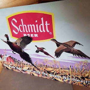 May include: Vintage Schmidt Beer sign featuring three pheasants in flight over a farm scene. The sign has a red and yellow logo with the text "Schmidt Beer" and the tagline "The Brew that Grew with the Great Northwest!"