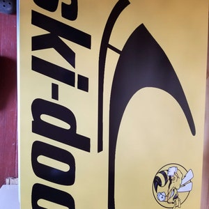 Puede incluir: Cartel rectangular amarillo con el texto negro "ski-doo" verticalmente a la izquierda y un gr&aacute;fico negro estilizado a la derecha. Un logotipo circular de una abeja de dibujos animados est&aacute; en la esquina inferior derecha.