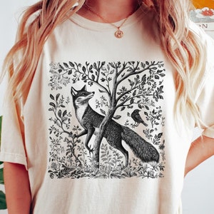 Peut inclure: Un t-shirt cr&egrave;me avec une illustration en noir et blanc d'un renard dans un d&eacute;cor de for&ecirc;t. Le renard est debout sur ses pattes arri&egrave;re et regarde vers la gauche. Un oiseau est perch&eacute; sur une branche au-dessus du renard.