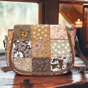 Może przedstawiać: Brązowa skórzana torba na ramię w stylu patchwork z kwiatowym i motylkowym wzorem. Torba ma długi regulowany pasek i zapięcie na klapę.