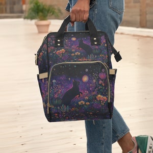 May include: A black backpack with a floral and starry pattern featuring black cats. The backpack has a zippered front pocket and adjustable straps.