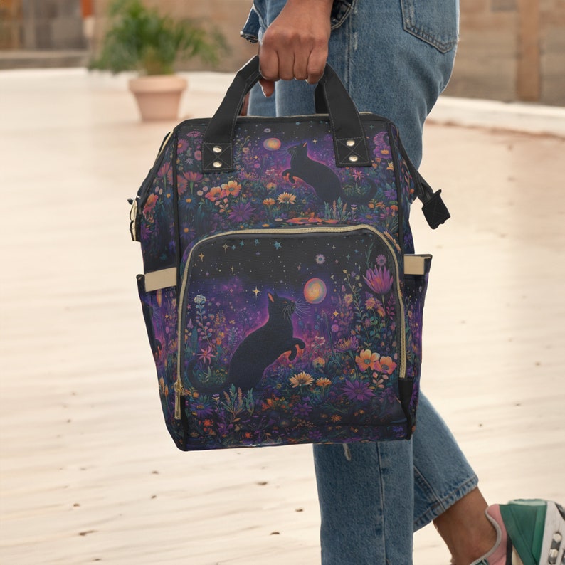 May include: A black backpack with a floral and starry pattern featuring black cats. The backpack has a zippered front pocket and adjustable straps.