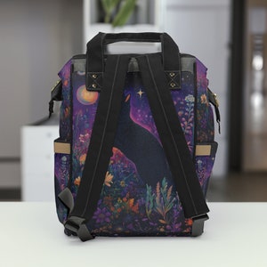 May include: A black backpack with adjustable straps features a colorful print of a black cat silhouetted against a purple and blue starry night sky. The print also includes a crescent moon and flowers.