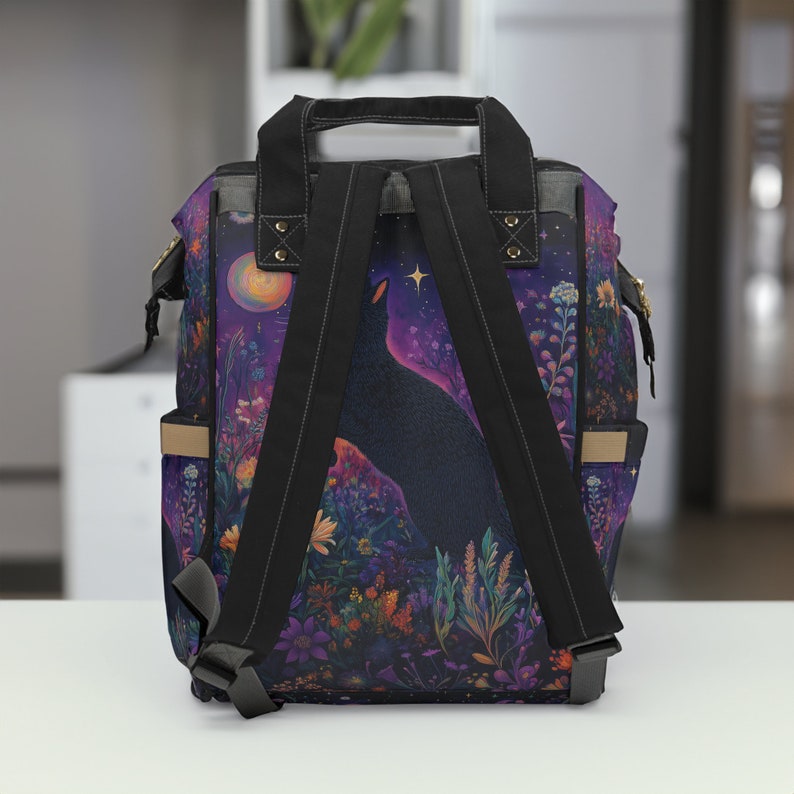 May include: A black backpack with adjustable straps features a colorful print of a black cat silhouetted against a purple and blue starry night sky. The print also includes a crescent moon and flowers.