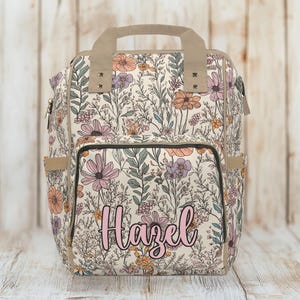 May include: A beige floral backpack with a black zipper and a pink name "Hazel" on the front.