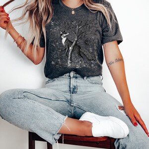 Peut inclure: Un t-shirt gris anthracite avec une illustration en noir et blanc d'un renard debout sur ses pattes arri&egrave;re devant un arbre. Le renard regarde vers la gauche. Le t-shirt est port&eacute; par une personne aux cheveux longs blonds et un jean bleu.