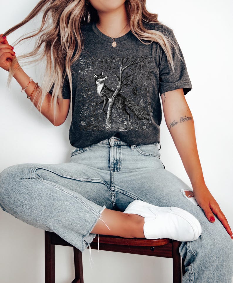 Peut inclure: Un t-shirt gris anthracite avec une illustration en noir et blanc d'un renard debout sur ses pattes arri&egrave;re devant un arbre. Le renard regarde vers la gauche. Le t-shirt est port&eacute; par une personne aux cheveux longs blonds et un jean bleu.