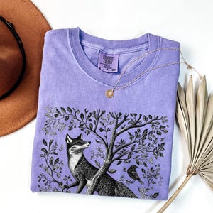 Peut inclure: Un t-shirt lavande avec une illustration en noir et blanc d'un renard dans une sc&egrave;ne de for&ecirc;t. Le t-shirt porte le texte "Comfort Colors" imprim&eacute; sur l'&eacute;tiquette.
