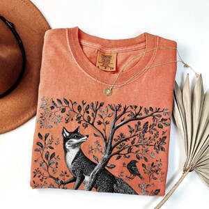 Peut inclure: Un t-shirt de couleur corail avec une illustration en noir et blanc d'un renard dans un arbre. Le t-shirt porte l'inscription "Comfort Colors" imprim&eacute;e sur le devant.