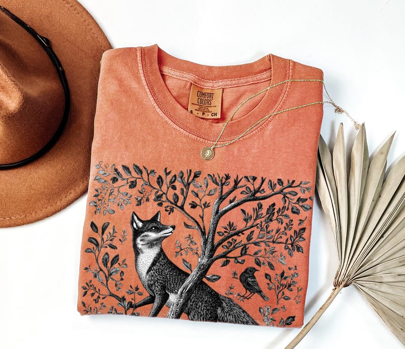 Peut inclure: Un t-shirt de couleur corail avec une illustration en noir et blanc d'un renard dans un arbre. Le t-shirt porte l'inscription "Comfort Colors" imprim&eacute;e sur le devant.